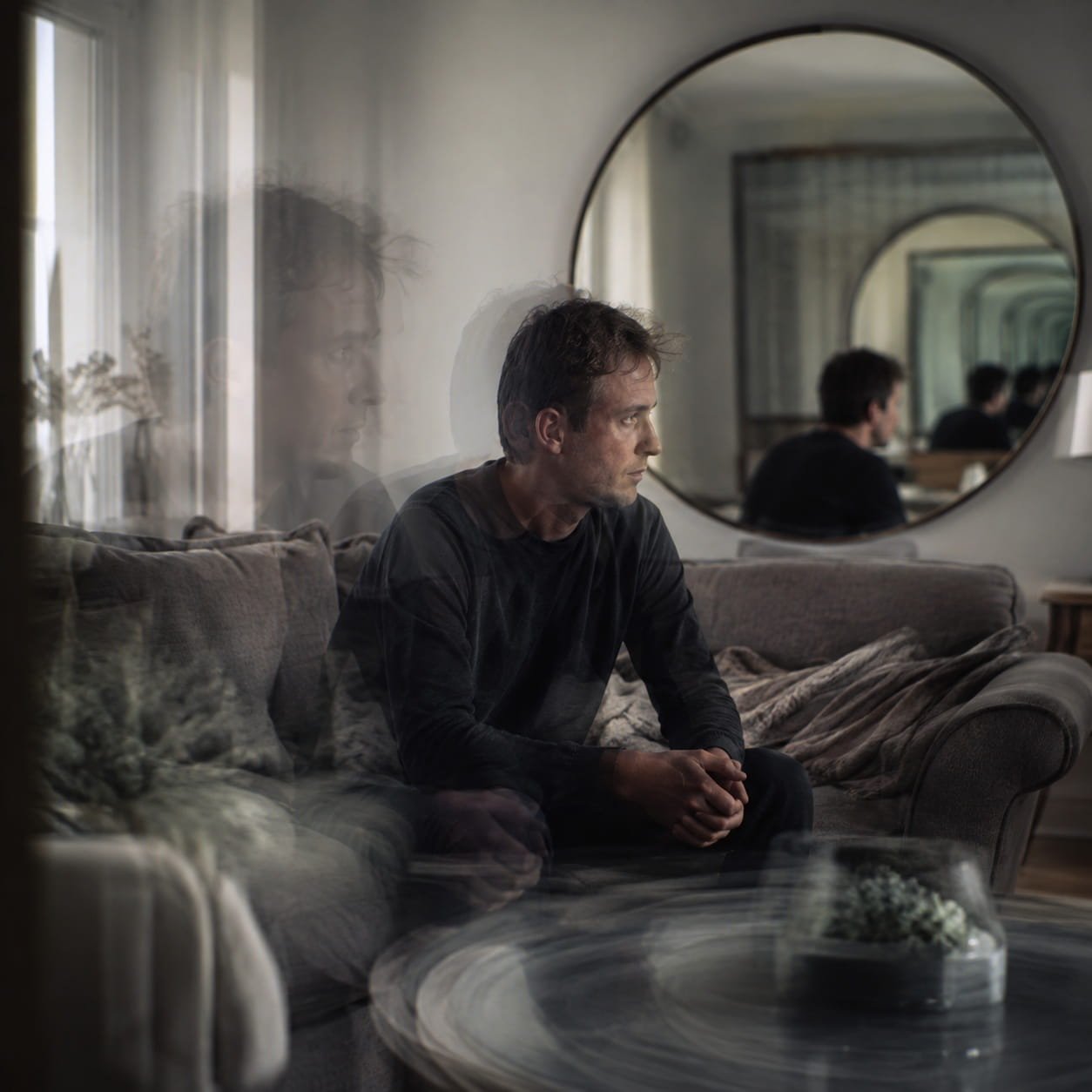 Existential OCD & Depersonalisation: Understanding 7 Vital Insights. Man sitting thoughtfully on a sofa surrounded by subtle reflections and double-exposure effects, representing existential OCD, depersonalisation and feelings of unreality.